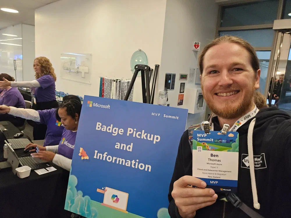 A person at a conference smiling and holding a badge and a sign that says &ldquo;Badge Pickup and Information&rdquo; at the MVP Summit.