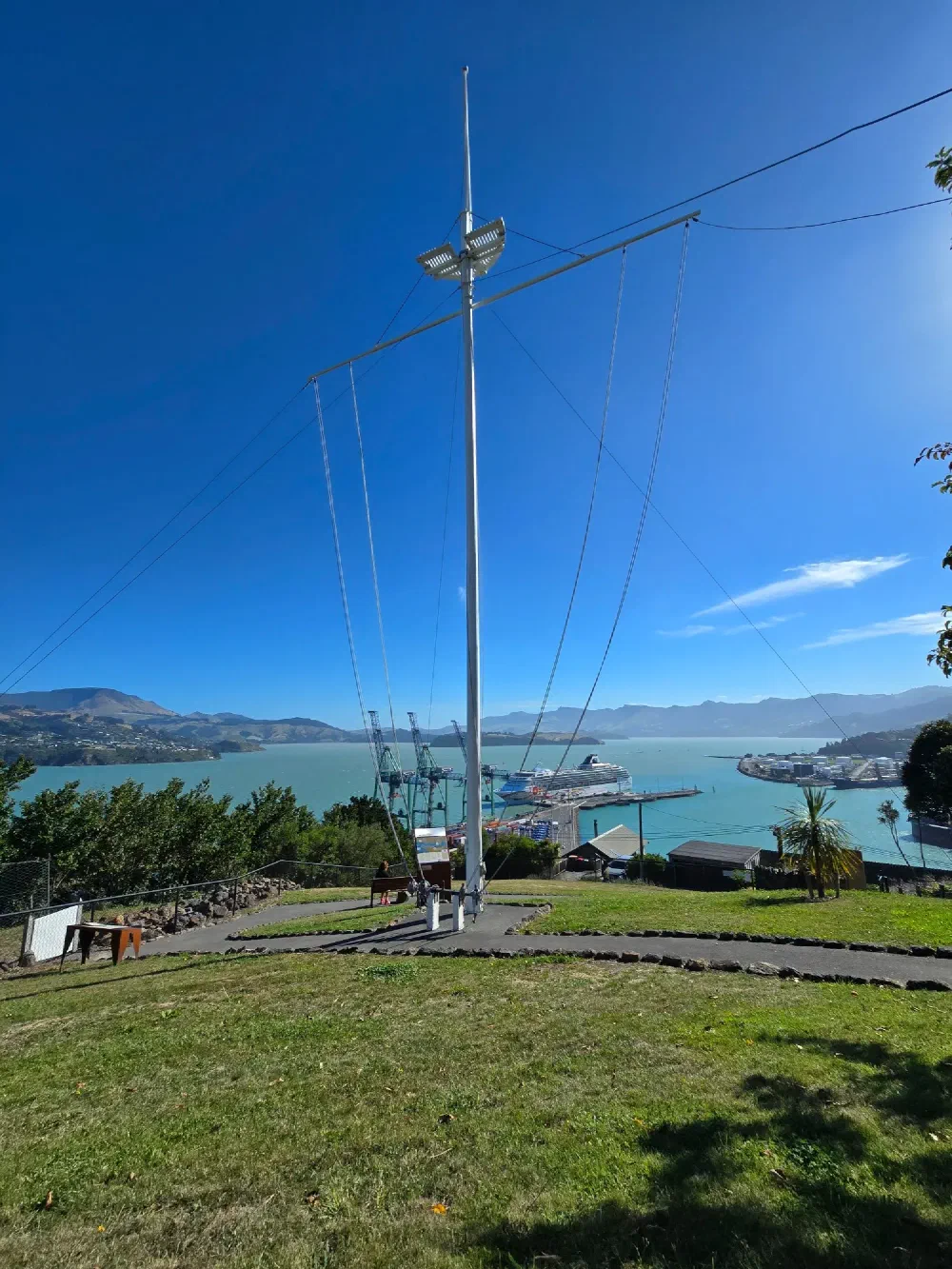 Tall ship mast on grassy hill overlooking a harbor with docks, ships, and surrounding hills under a clear blue sky.