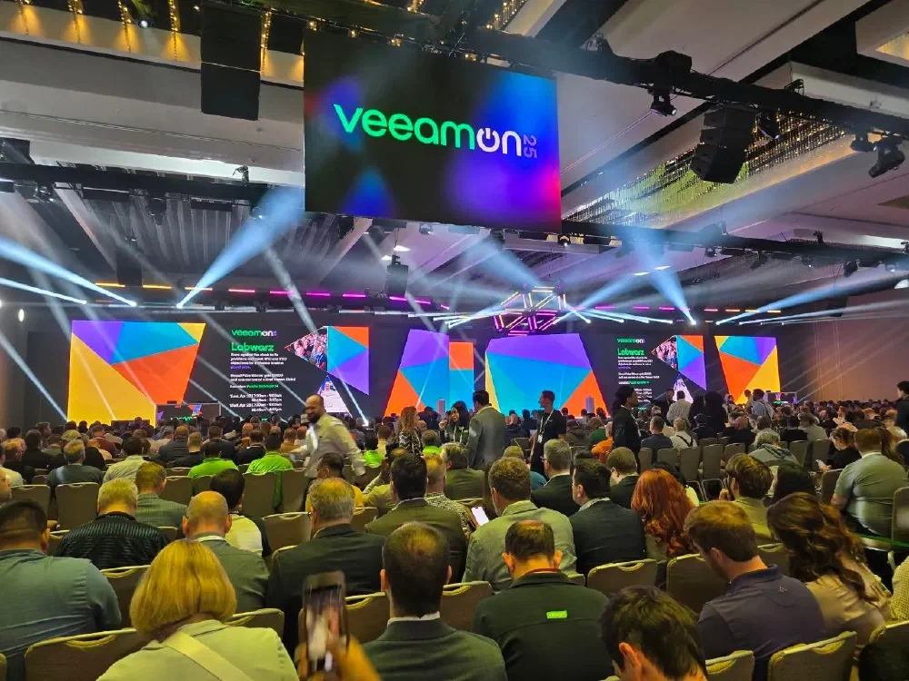 A view from the back of a large conference room filled with people watching presentations on multiple screens on a stage at a VeeamON event.