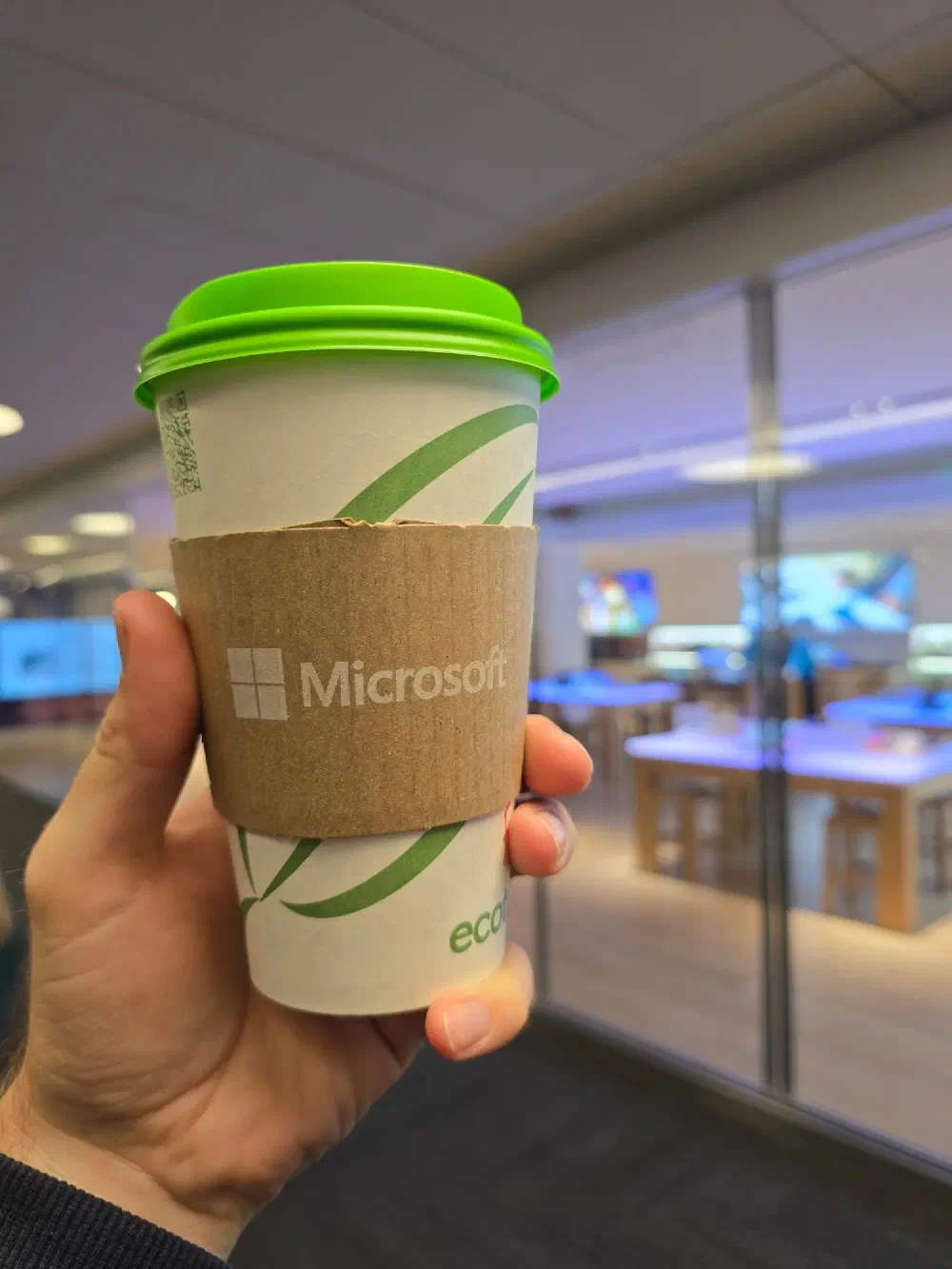 A hand holding a paper coffee cup with a green lid and a cardboard sleeve with the Microsoft logo, in a blurry indoor setting.
