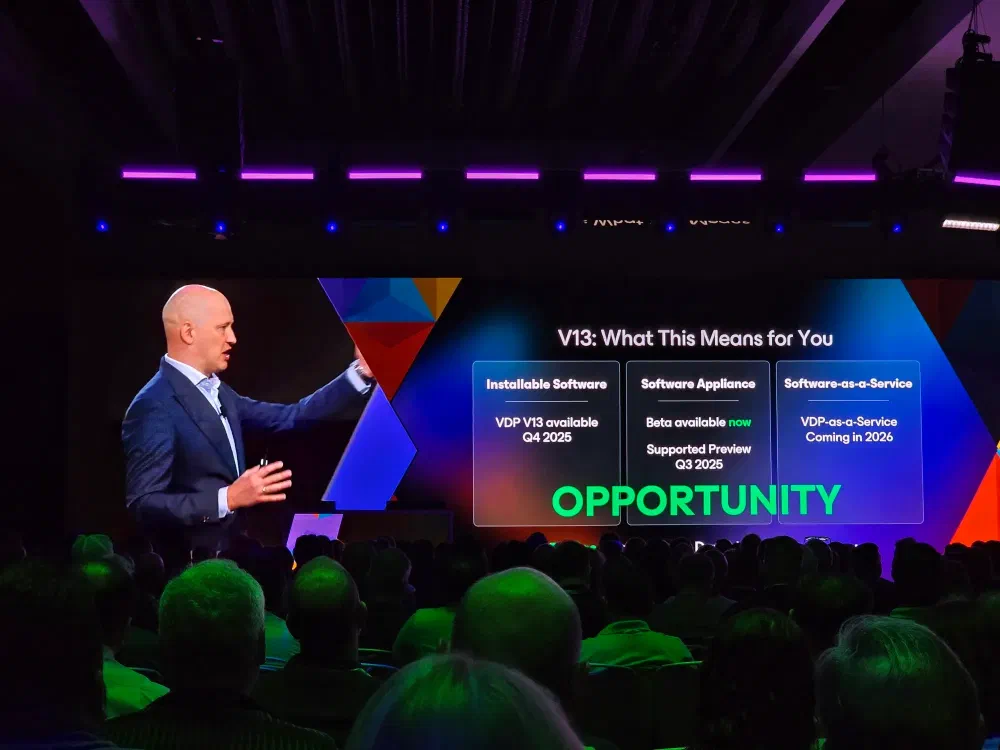 A speaker on stage at a conference points to a large screen displaying &ldquo;V13: What This Means for You&rdquo; and details about software availability and &ldquo;OPPORTUNITY&rdquo;. An audience is visible in the foreground.