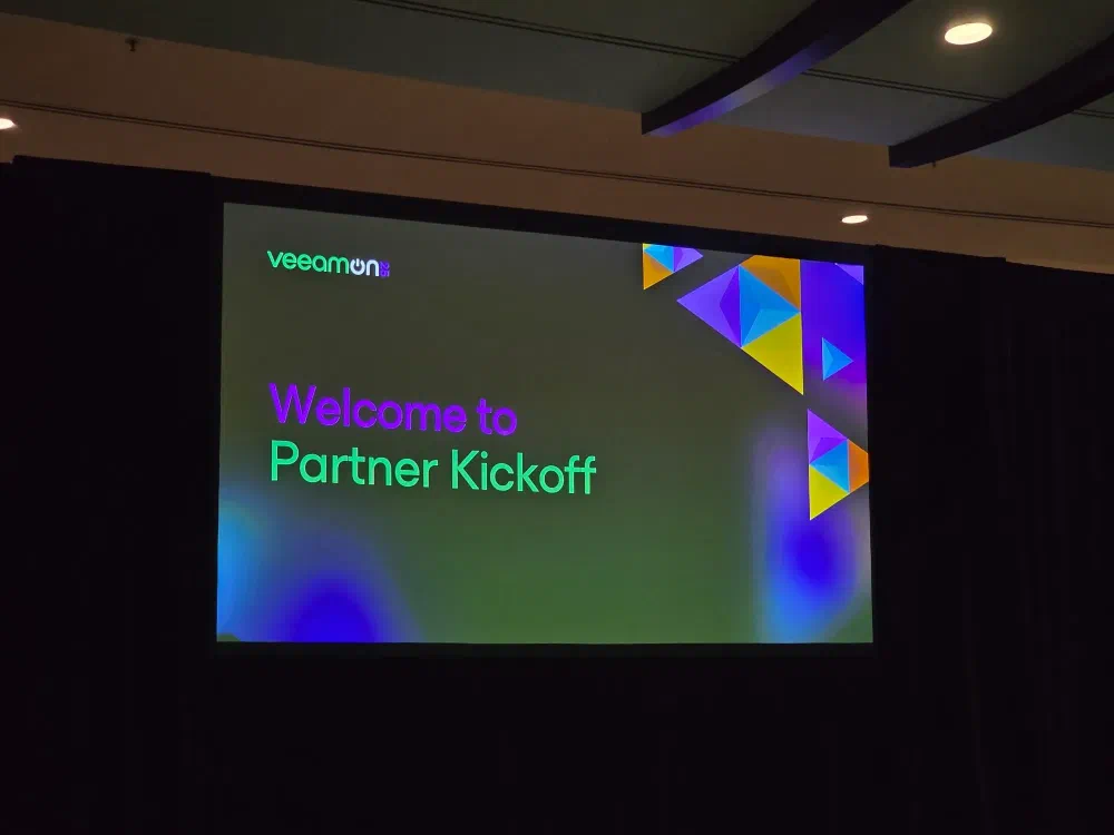 A screen displays &ldquo;veeamon Welcome to Partner Kickoff&rdquo; with colorful abstract shapes in the background.