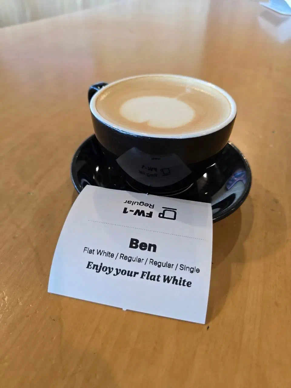 A close-up of a flat white coffee with latte art, sitting on a table next to a label showing the name Ben and details of the coffee order.