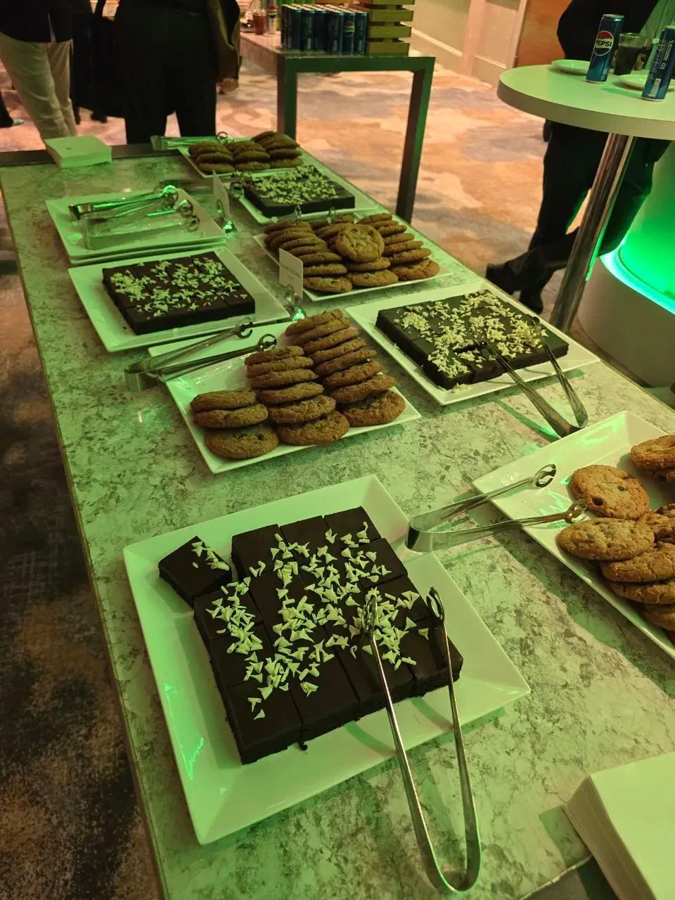 A table is filled with plates of desserts, including chocolate brownies with white sprinkles and stacks of cookies, with serving tongs on the plates.