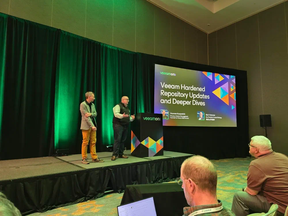 Two speakers are on a stage at a conference, presenting in front of a screen that displays “VeeamON: Veeam Hardened Repository Updates and Deeper Dives” and their names and titles. An audience is visible in the foreground.