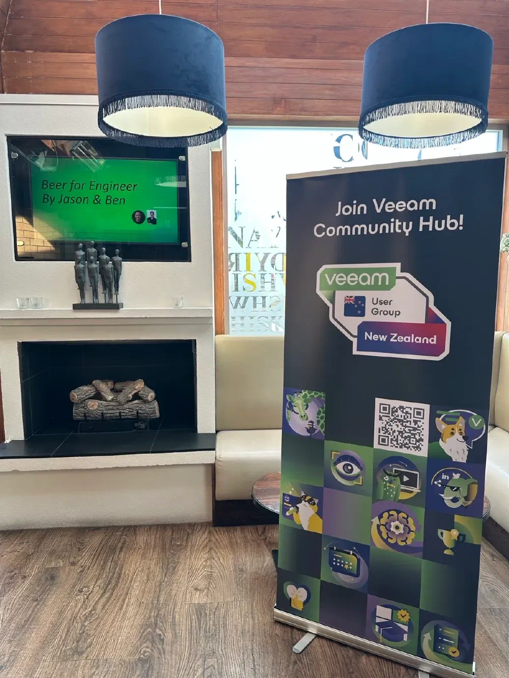 A promotional banner for &ldquo;Veeam User Group New Zealand&rdquo; stands next to a fireplace with a TV displaying &ldquo;Beer for Engineer By Jason & Ben&rdquo;.

